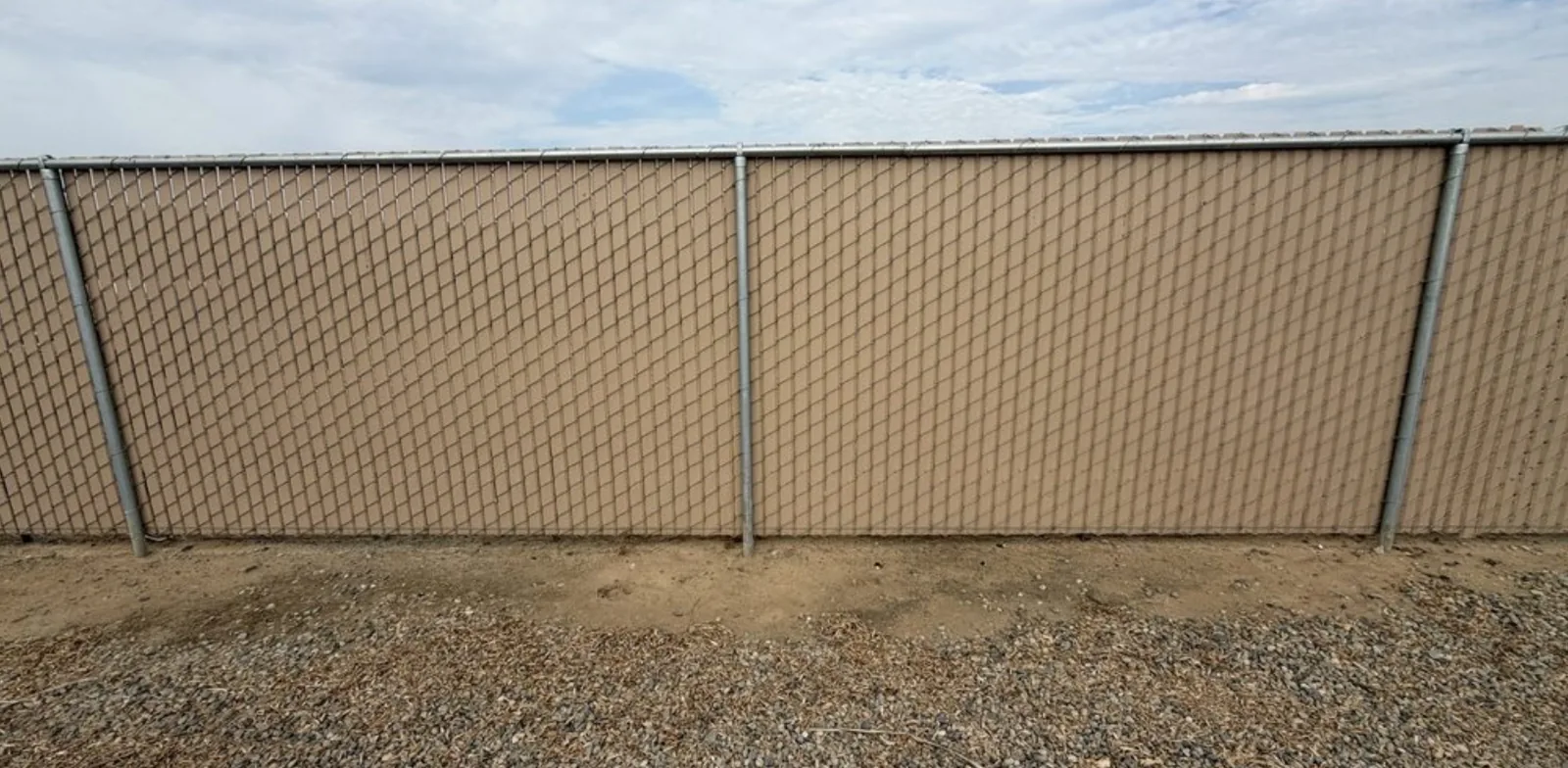 Tan privacy-slat chain-link fence built by AR Fence along a property line, with galvanized line posts and top rail and a gravel strip at the base
